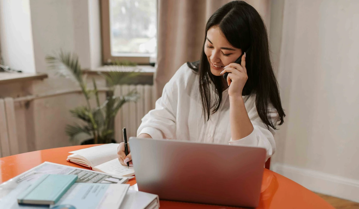 Young woman talking on phone while writing notes at a desk with a laptop and documents.