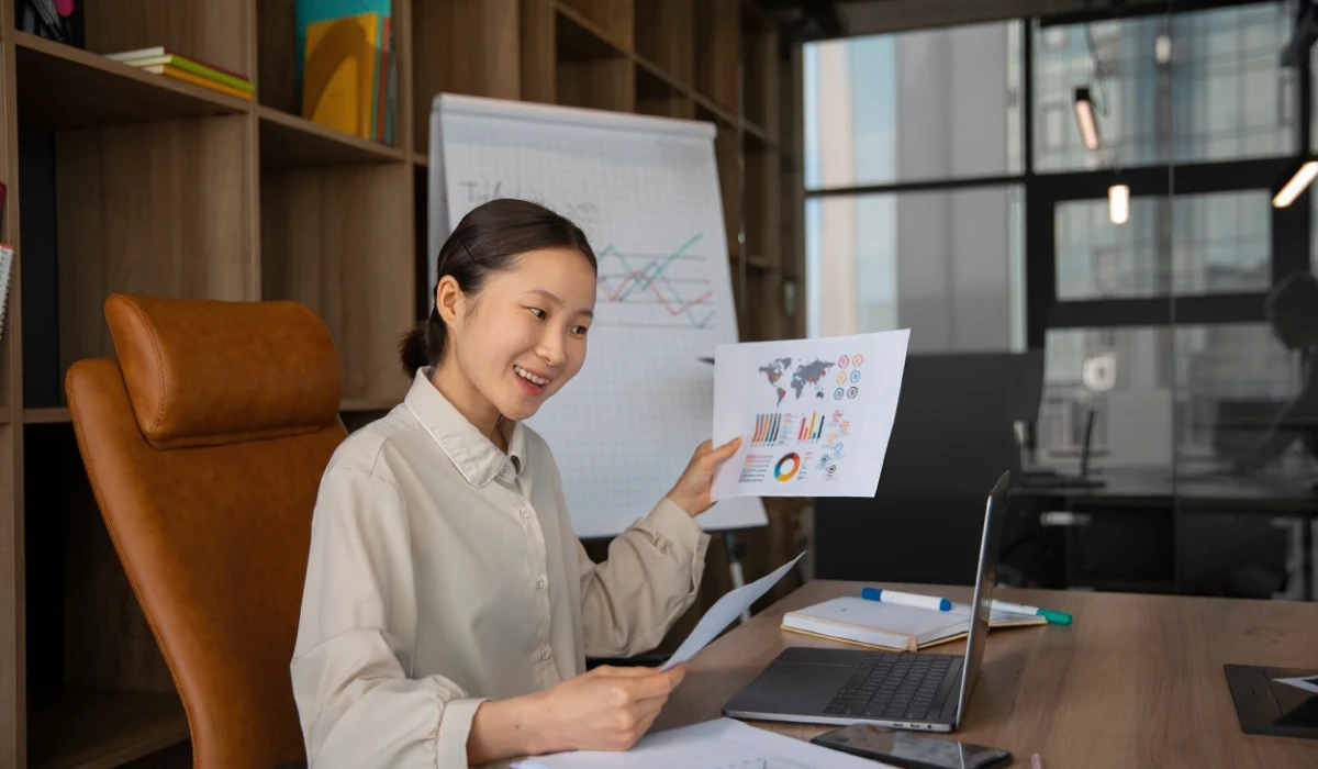 Smiling businesswoman holding charts and graphs while working on a laptop in a modern office.