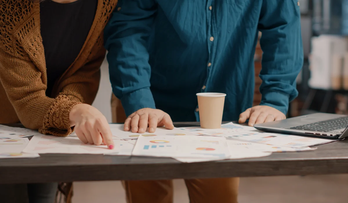 Two people reviewing and pointing at charts and graphs spread on a table with a laptop and paper cup nearby.
