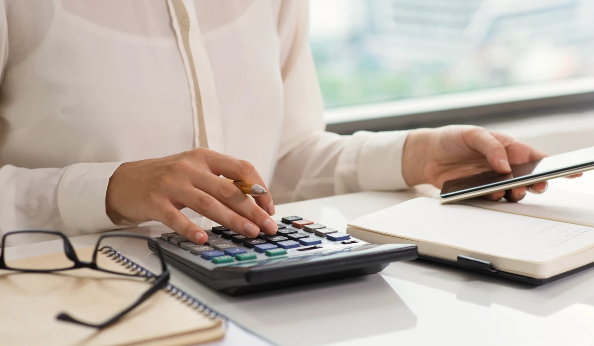 Person using a calculator with one hand and holding a smartphone over an open notebook on a desk with glasses nearby.