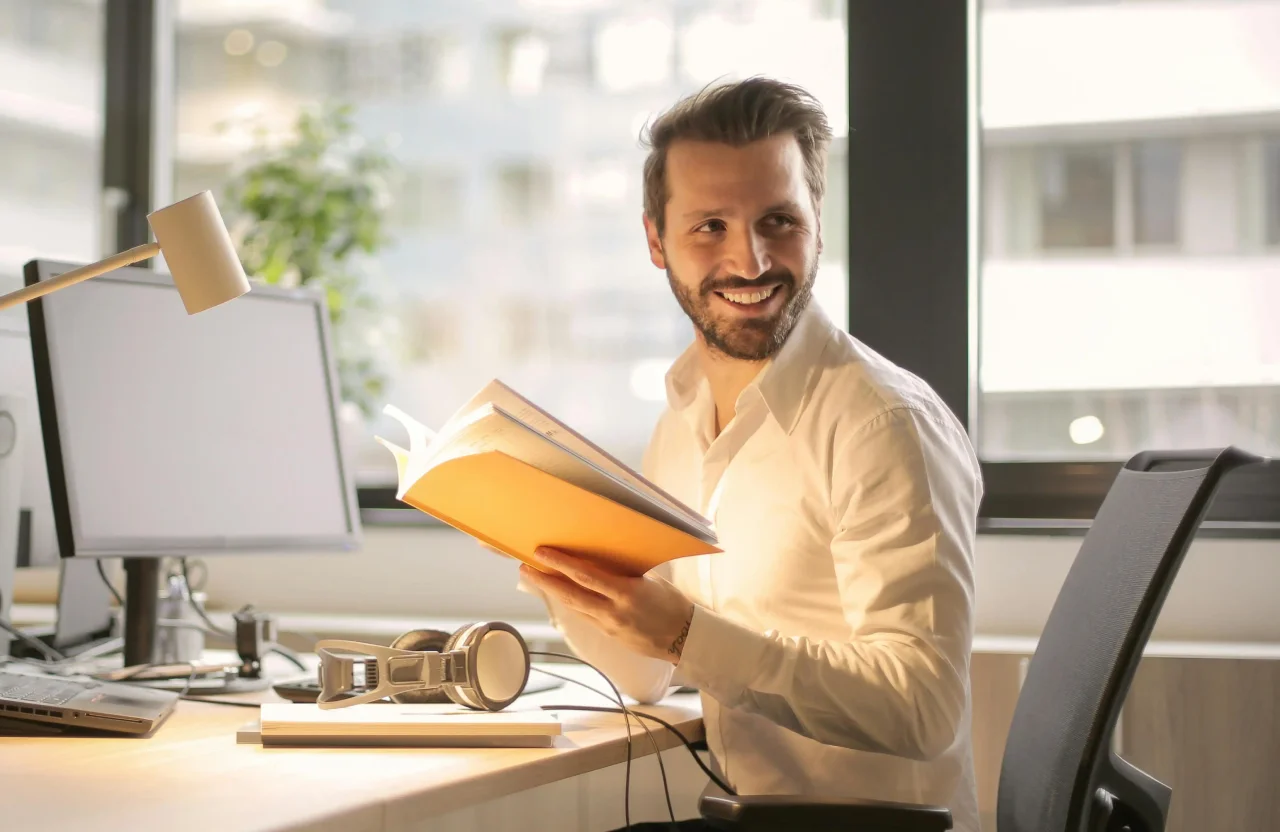 Smiling man in white shirt sitting at a desk holding an open book in a bright office.