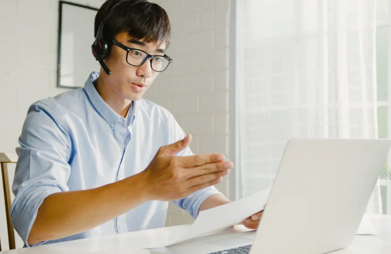Young man wearing glasses and a headset, gesturing while talking during a video call at a desk with a laptop.