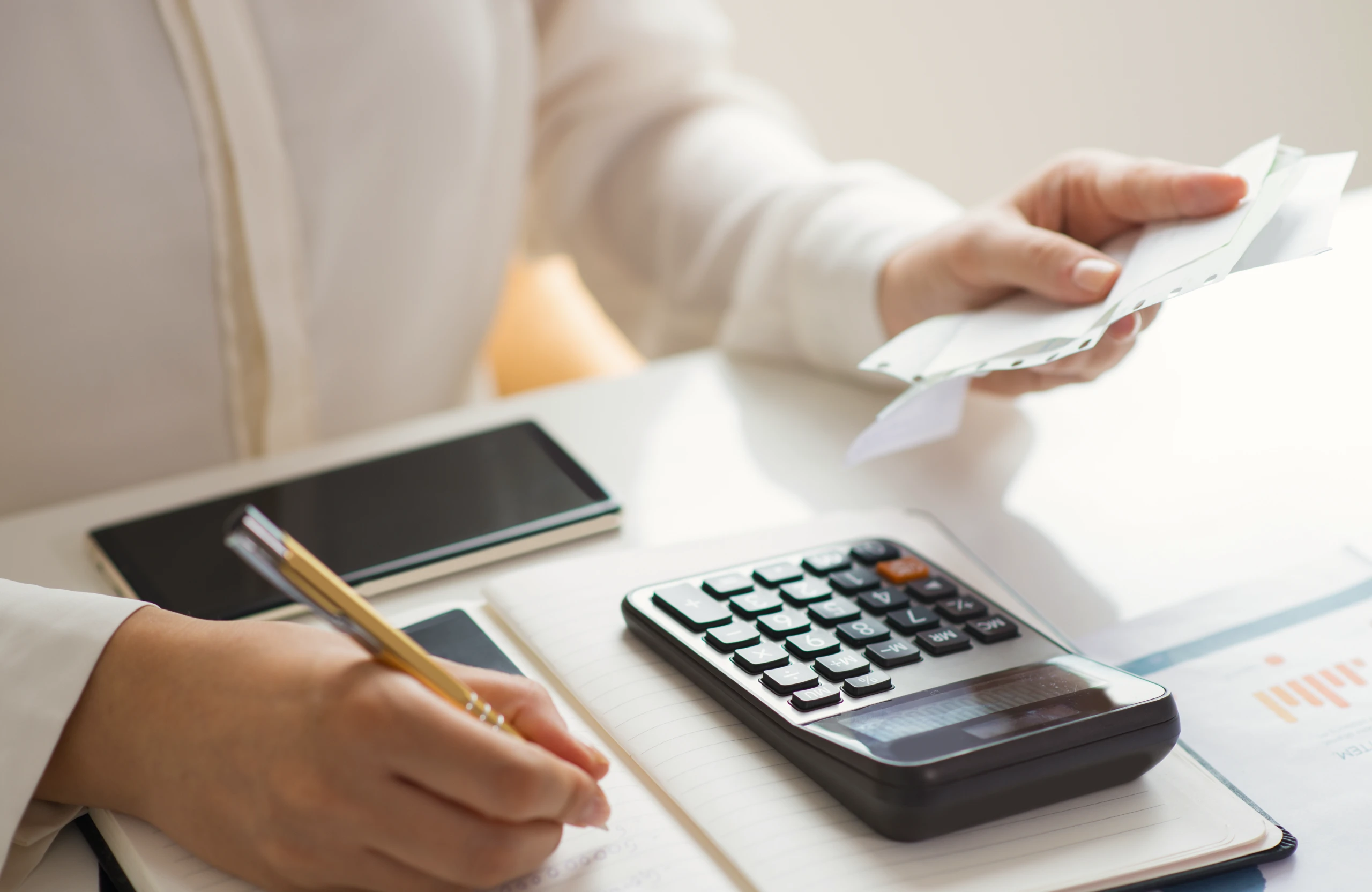 Person using a calculator and writing notes while holding receipts at a desk with a smartphone and documents.