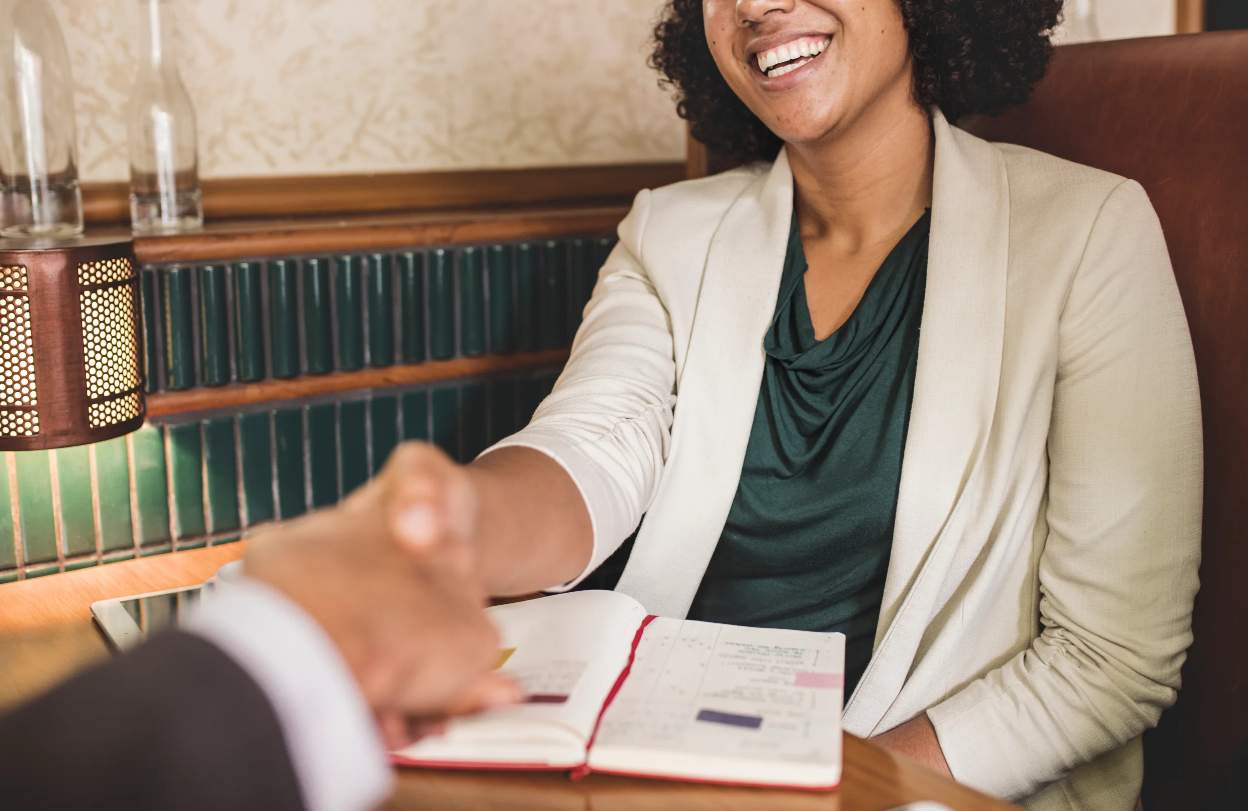 Smiling woman in a cream blazer shaking hands across a table with an open planner.