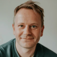 Portrait of a smiling man with short light brown hair wearing a dark green shirt against a plain background.