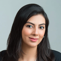 Headshot of a smiling woman with long black hair wearing a black blazer and light pink lipstick, against a plain light gray background.