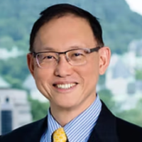 Smiling man wearing glasses, a dark suit, striped shirt, and yellow tie, with a blurred outdoor background.