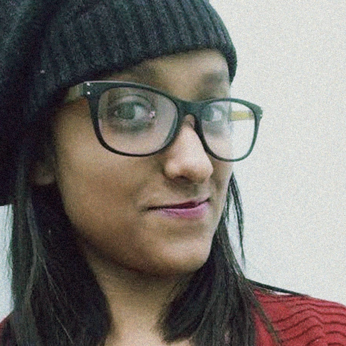 Close-up of a woman with long dark hair wearing black glasses and a black knit hat, smiling slightly, with a red top.