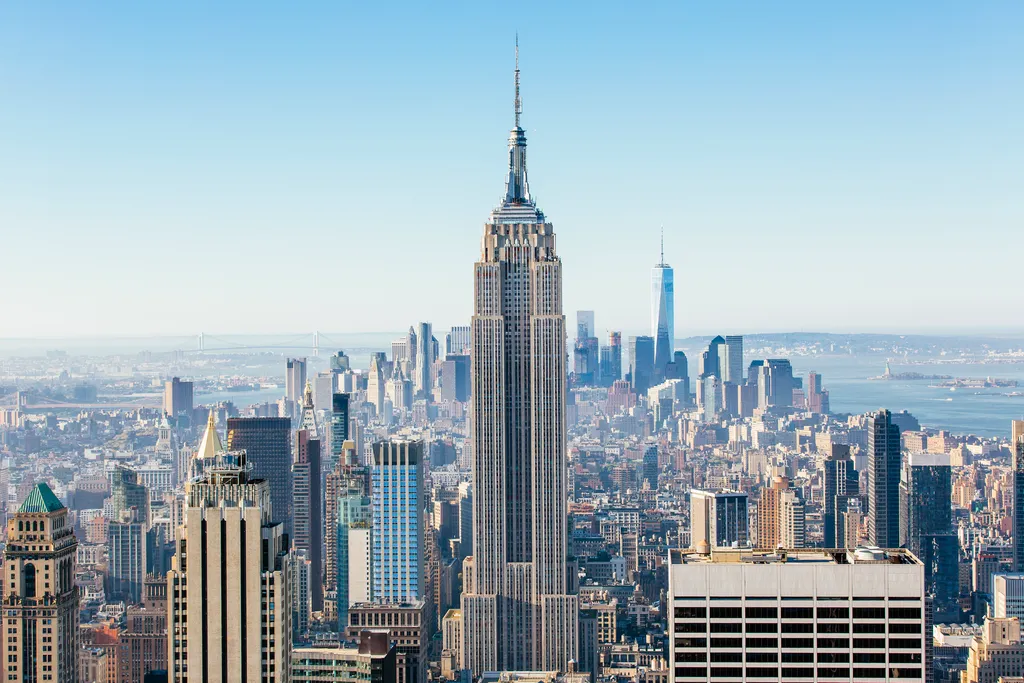 View of New York City skyline with the Empire State Building centered and One World Trade Center visible in the background under a clear blue sky.