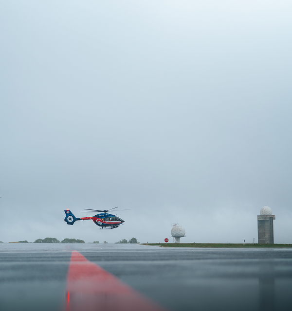 Blue and red helicopter hovering over a wet runway under an overcast sky near radar towers.