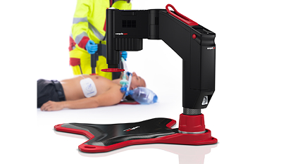 Robotic device performing CPR on a mannequin with a person in high-visibility clothing in the background.