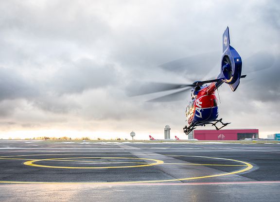Blue and red medical helicopter lifting off from a helipad under a cloudy sky.