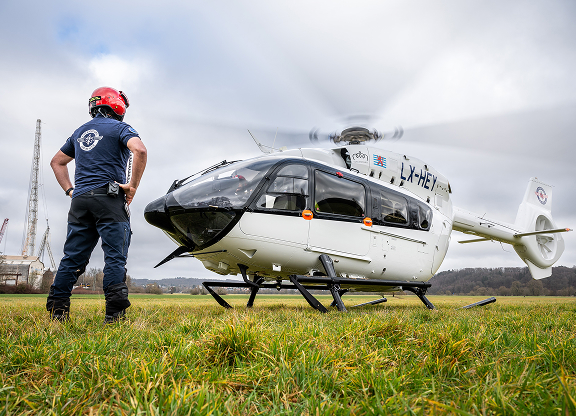 Rescue worker in a red helmet standing on grass near a white helicopter with spinning rotor blades.