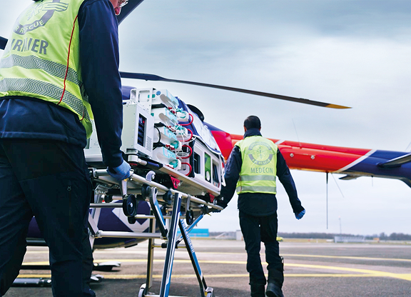 Rescue workers transporting medical equipment on a stretcher near a red and blue helicopter on a helipad.
