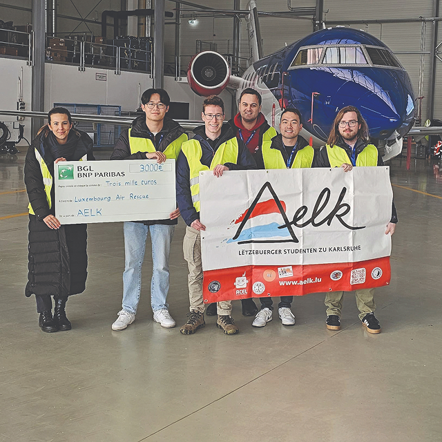 Group of six people standing in an airplane hangar holding a large check and a banner for AELK in front of a blue plane.