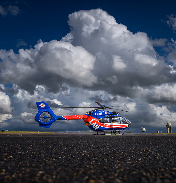 Blue and red rescue helicopter with registration LX-HSL on runway under dramatic cloudy sky.