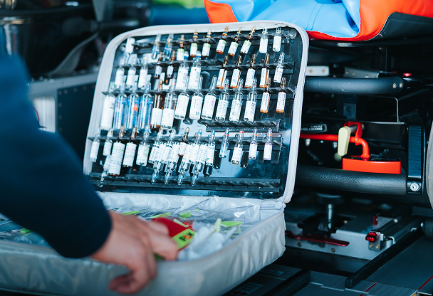 Open emergency medical kit with multiple vials and ampoules inside a vehicle, a hand reaching towards it.