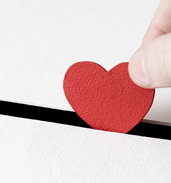 Hand placing a red textured heart shape into a narrow black slot on a white textured surface.