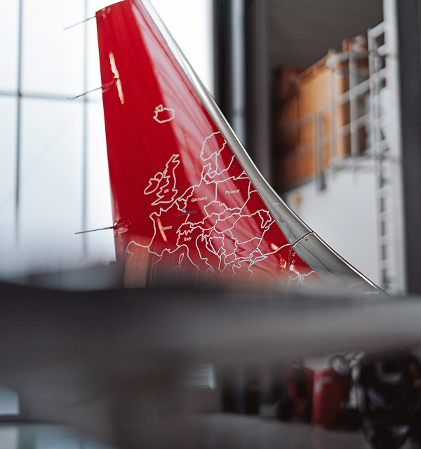 Close-up of a red airplane tail fin with a white outline map of Europe showing country names including Germany, Poland, and Italy, inside a hangar.