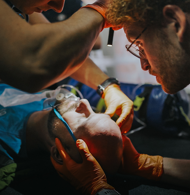 Medical personnel providing emergency care to a bald man wearing oxygen mask, with one person holding his head and another closely monitoring.
