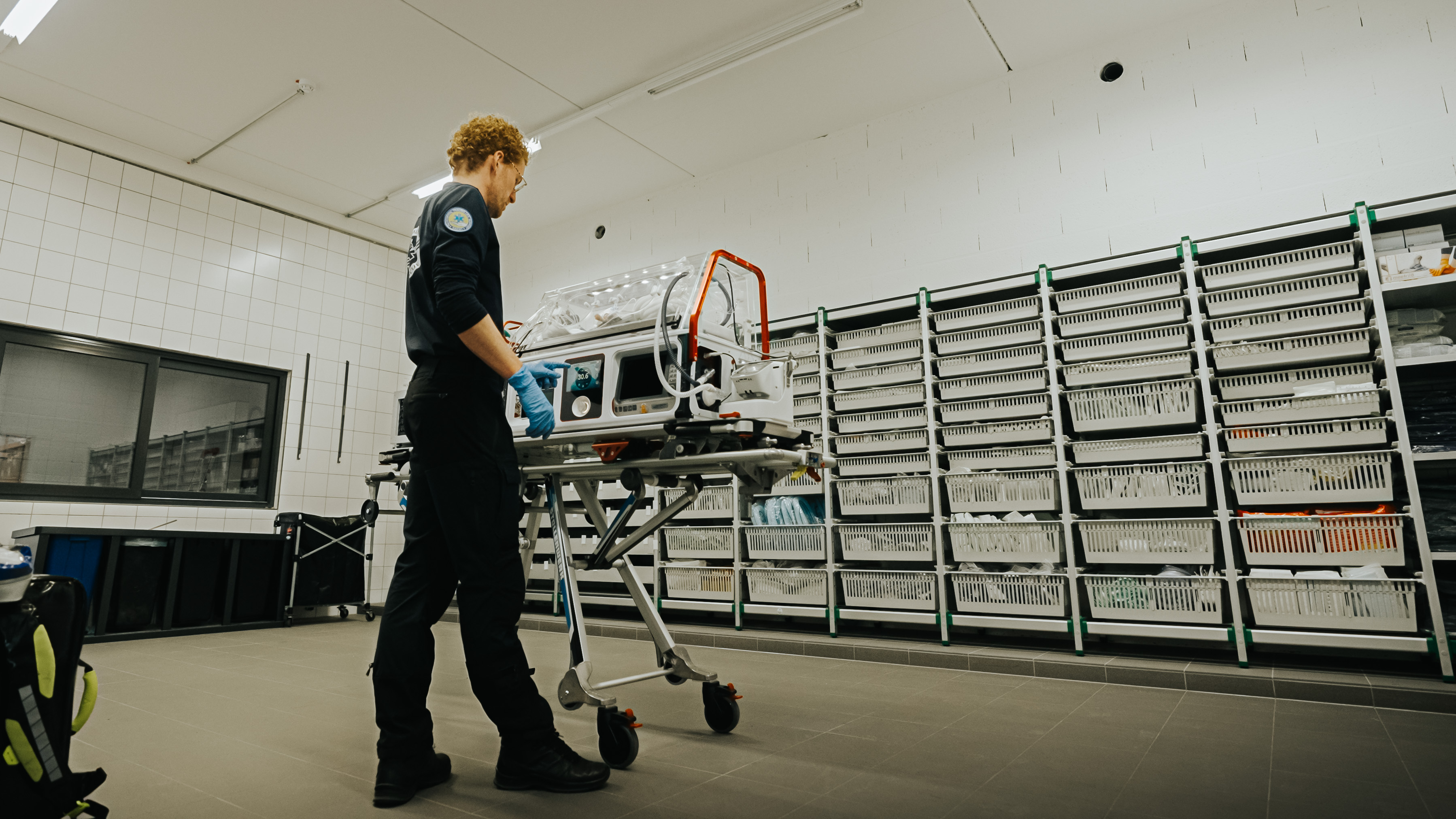 Medical professional adjusting neonatal incubator on wheeled stretcher in a storage room.