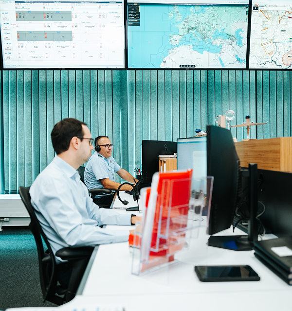 Two men working at desks with multiple computer screens in a control room with large monitors displaying maps and data on the wall.