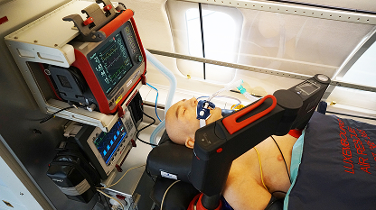 Medical mannequin lying on a stretcher inside an aircraft with monitoring equipment and a robotic arm positioned nearby.