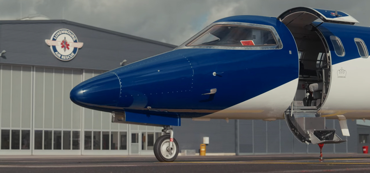 Close-up of a blue and white airplane nose with its door open, parked in front of a Luxembourg Air Rescue hangar.