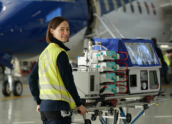 Smiling airport ground staff in a yellow safety vest standing beside medical equipment on a trolley in front of an airplane.
