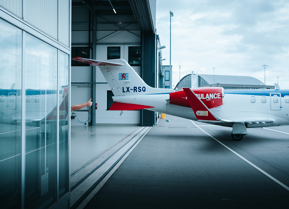 Tail section of a white and red ambulance airplane parked outside a hangar with a person giving a thumbs-up from inside the hangar.