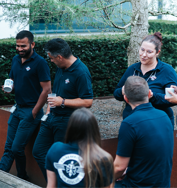 Five people wearing dark polo shirts with a medical logo, casually talking outdoors near a tree.