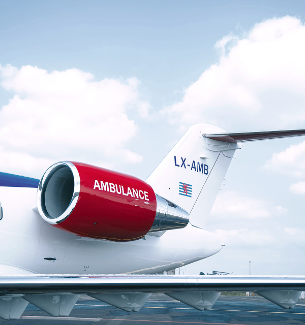 Close-up of a white airplane tail with a red engine marked 'AMBULANCE' and the registration LX-AMB against a partly cloudy sky.