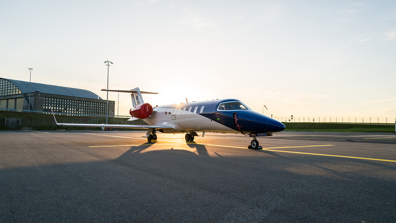 Private jet with a blue and white body and red engine covers parked on tarmac at sunset.