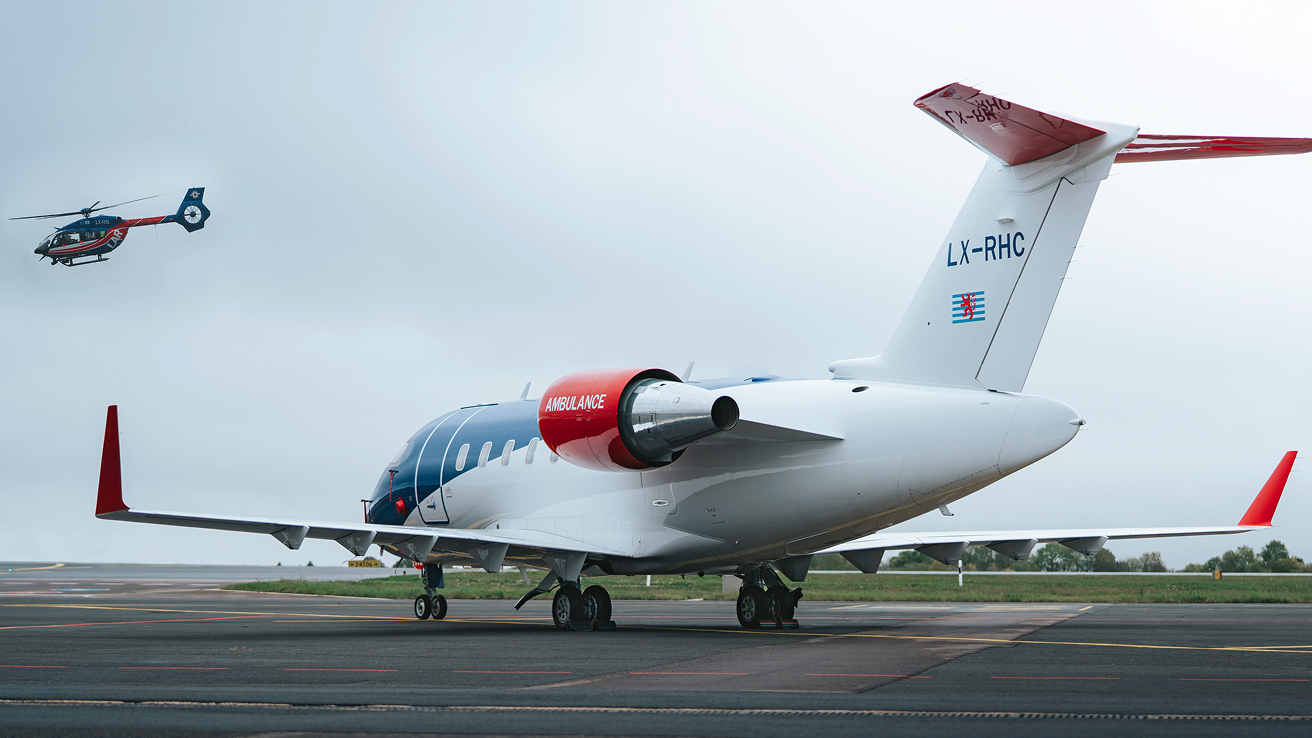 White and red medical ambulance jet parked on an airport runway with a blue and red helicopter flying in the cloudy sky.