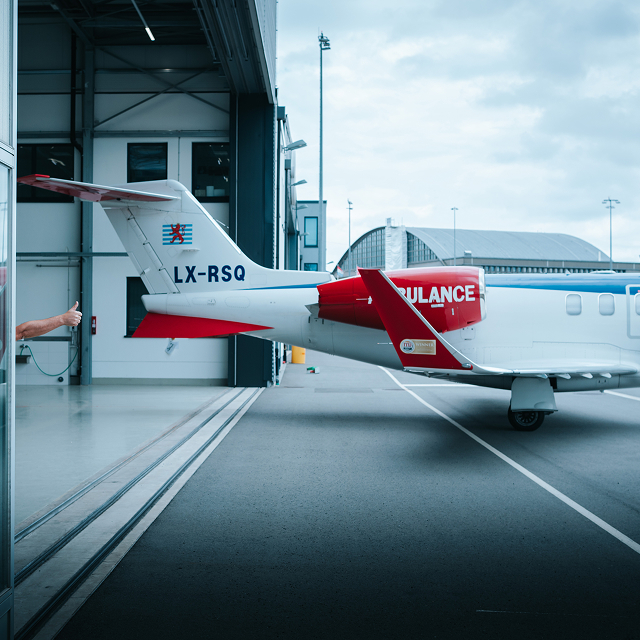 Rear view of a white and red ambulance jet parked partially inside an airport hangar, with a hand giving a thumbs-up from inside the hangar.