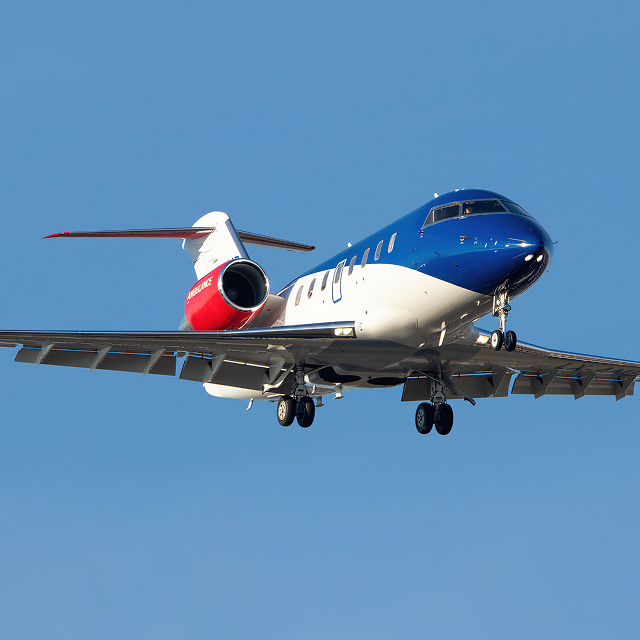 Blue and white private jet with red engine nacelle landing with wheels down against a clear blue sky.