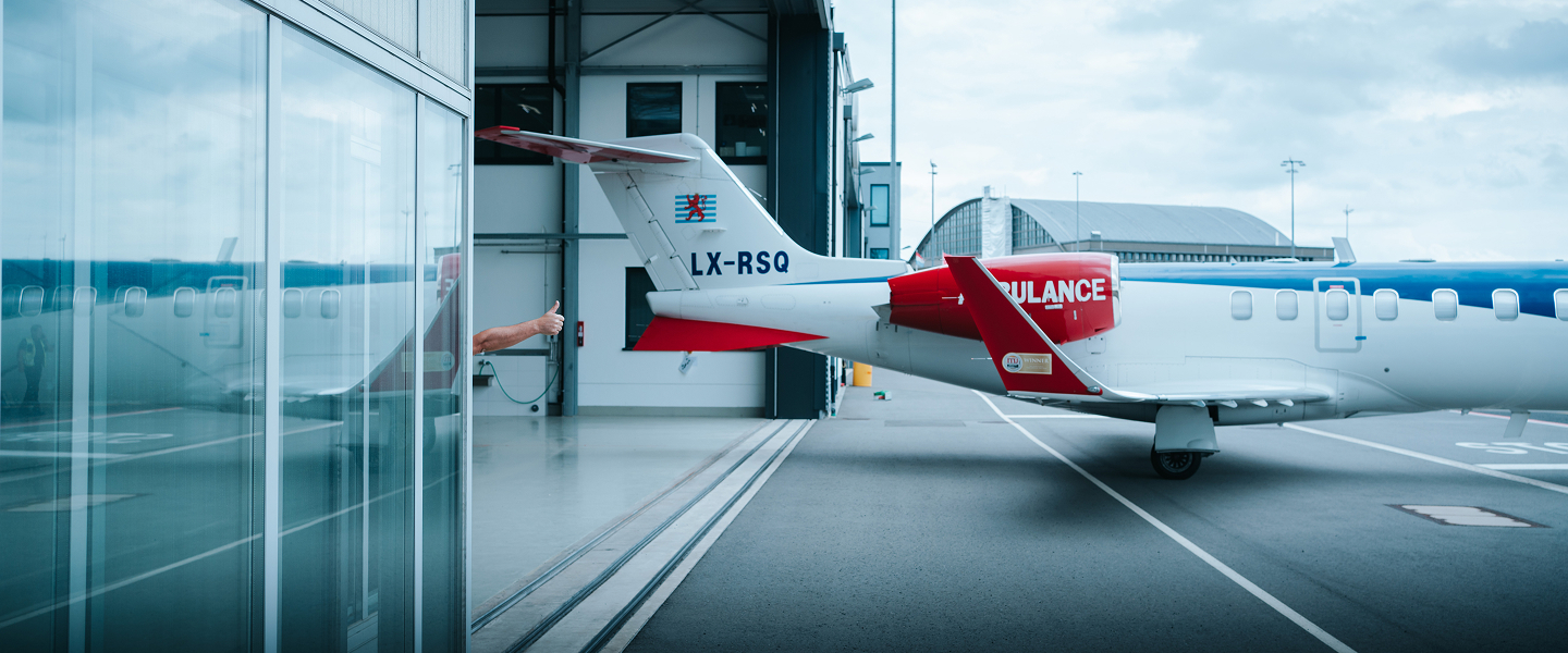 Tail section of a white and red ambulance aircraft parked by an airport hangar, with a person giving a thumbs up behind it.