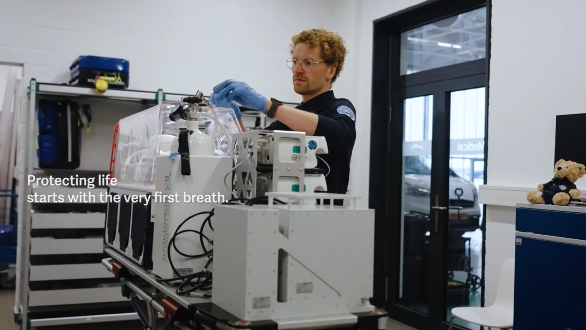 Technician wearing gloves adjusting equipment in a laboratory with medical device and teddy bear on a blue cabinet nearby.