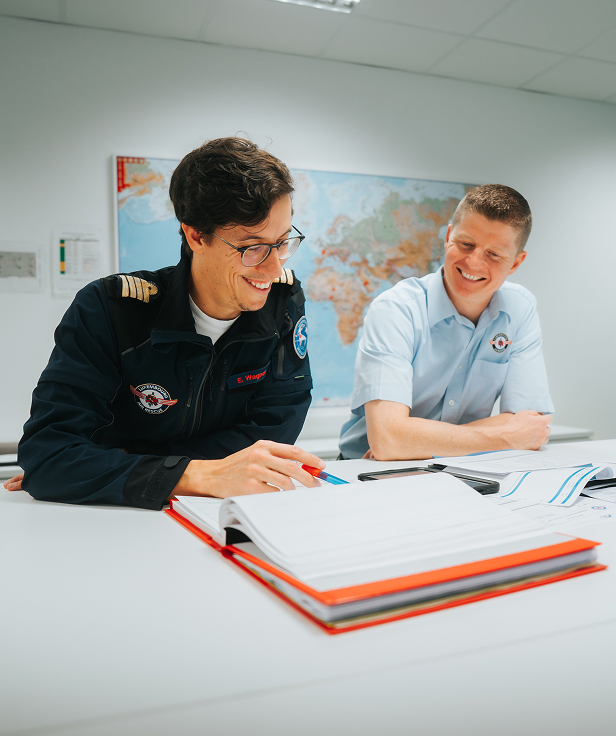 Two men in uniforms smiling and reviewing documents at a table with a world map in the background.