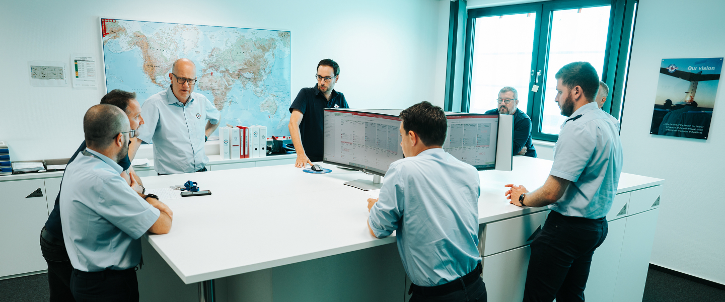 Eight men in light blue shirts and one in a dark shirt gathered around a white table with dual monitors, in an office with a world map and window.