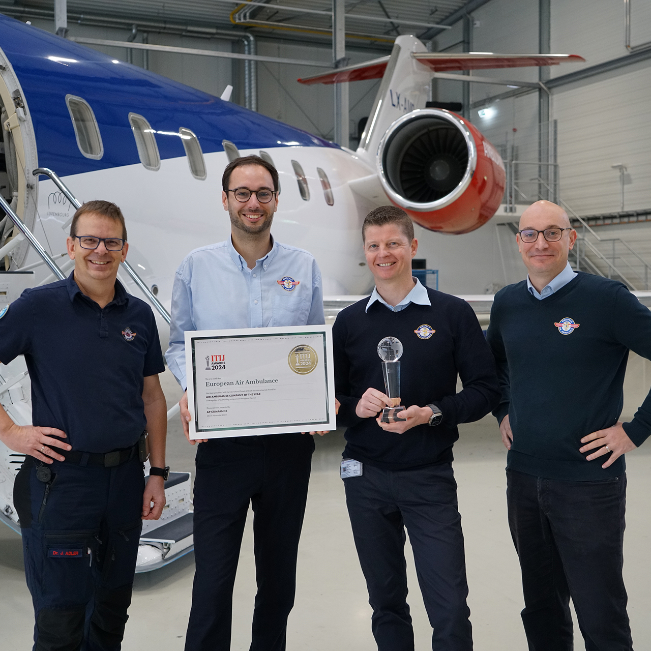 Four men standing in front of a private jet, two holding an award plaque and a trophy, smiling at the camera.
