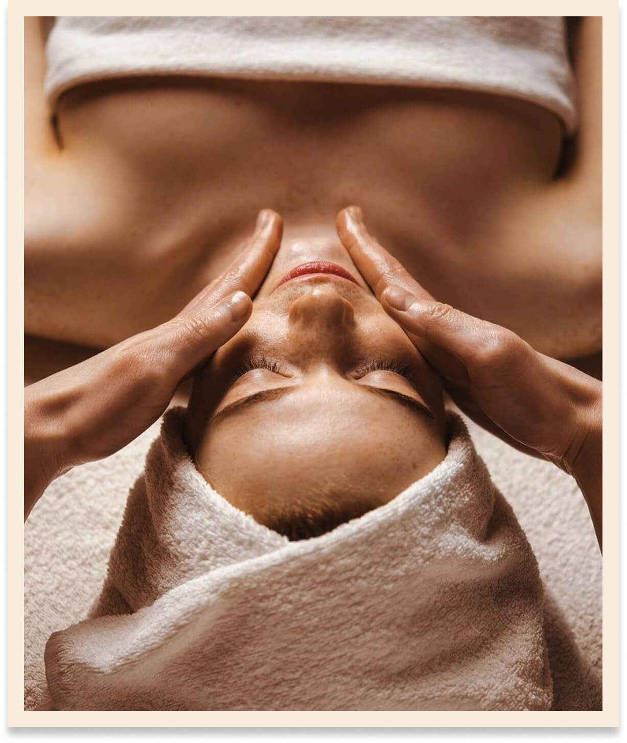 Person receiving a facial massage with hands gently pressing on both sides of the face, head wrapped in a towel.