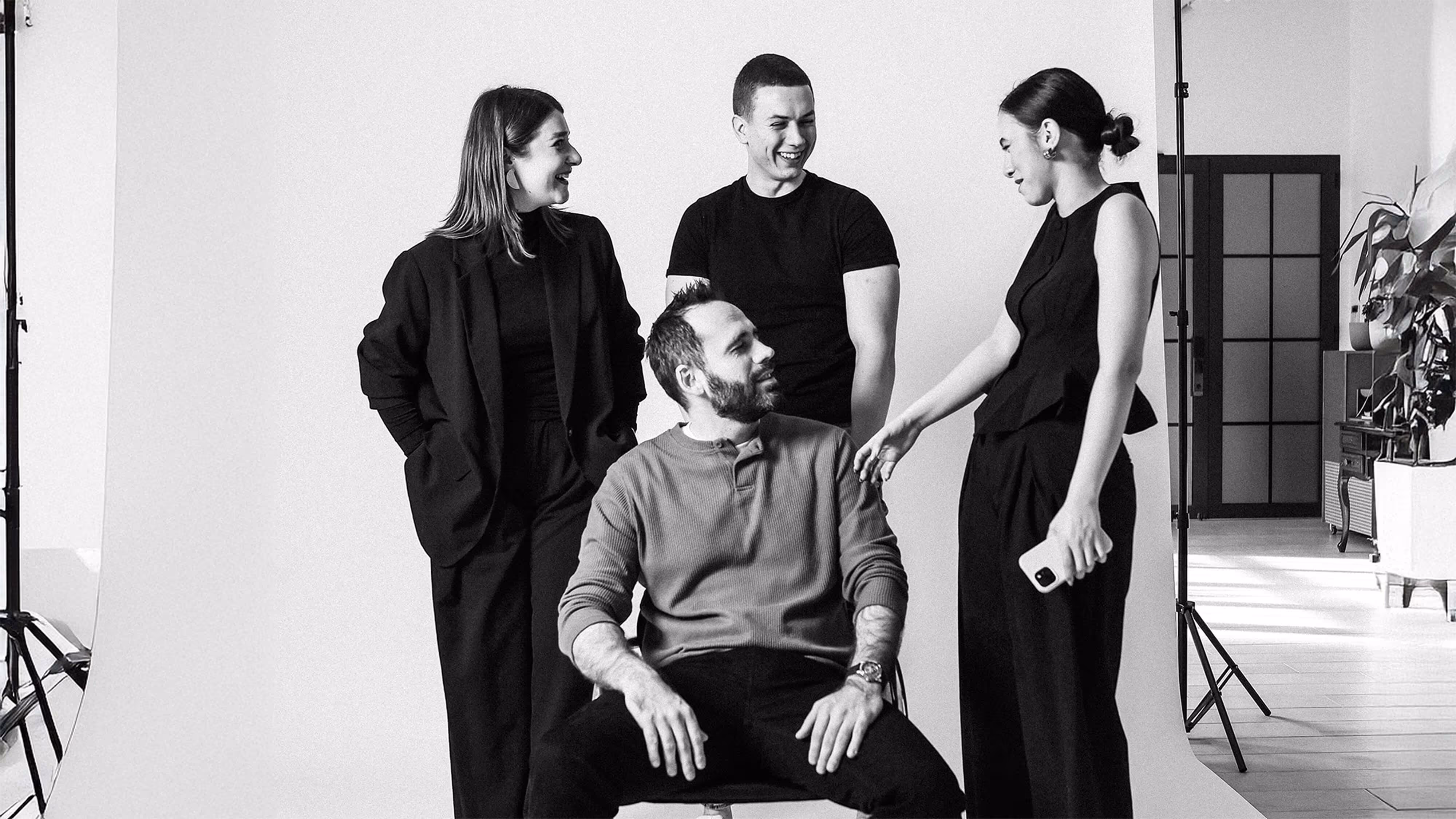 Seven diverse young adults sitting and standing around a sofa in a modern room, smiling and looking at the camera.