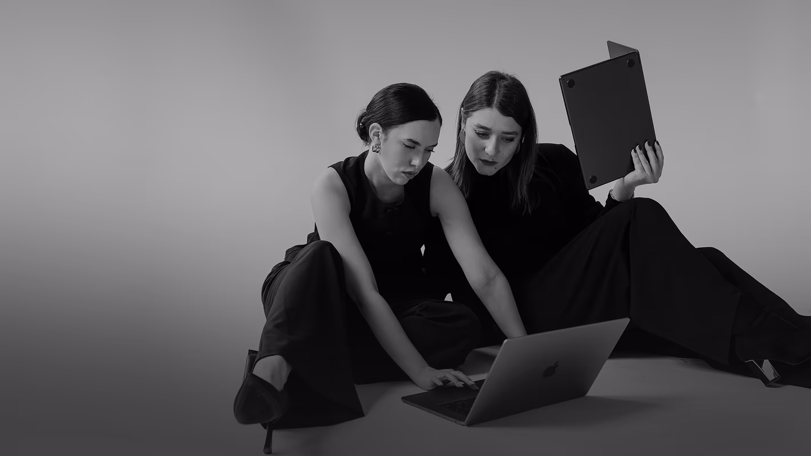 Two women sitting on the floor working on a laptop together, one holding a tablet.