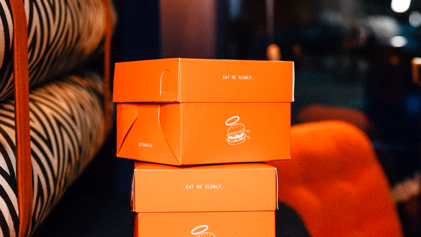 Two bright orange burger boxes stacked, each with 'EAT ME SLOWLY' text and burger icon with a halo.