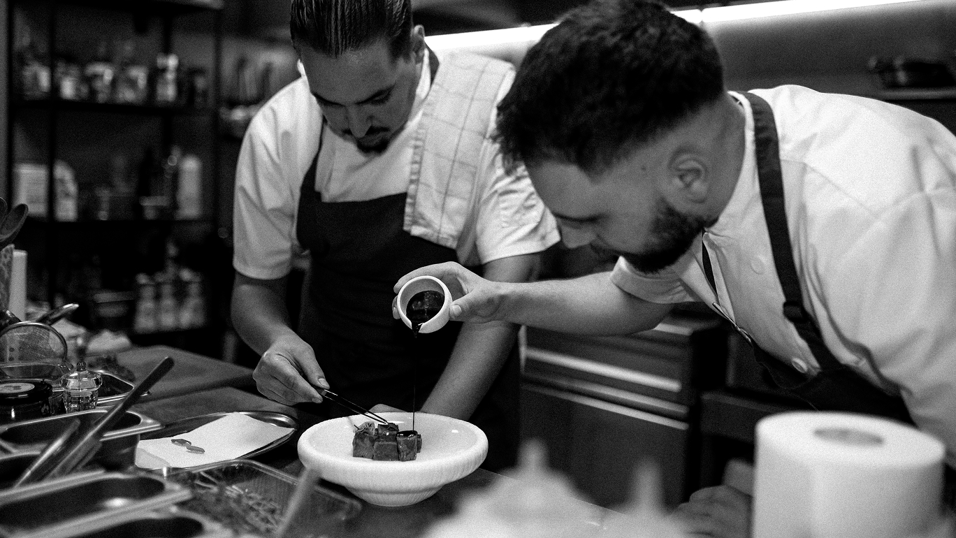 Two chefs in apron plating a gourmet dish in a professional kitchen.