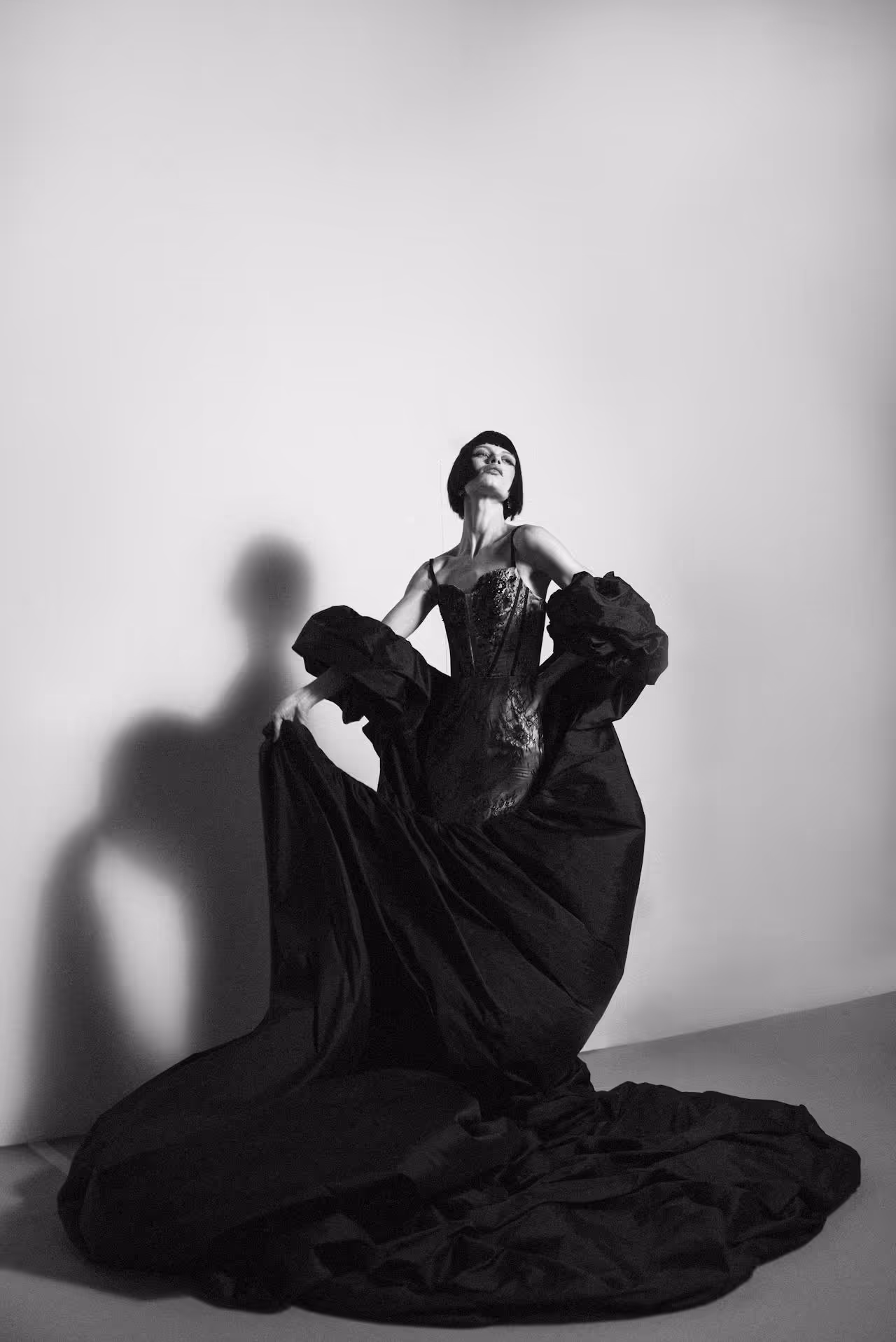 Black and white photo of a woman striking a dramatic pose in a voluminous black gown with puffed sleeves and a short bob haircut.