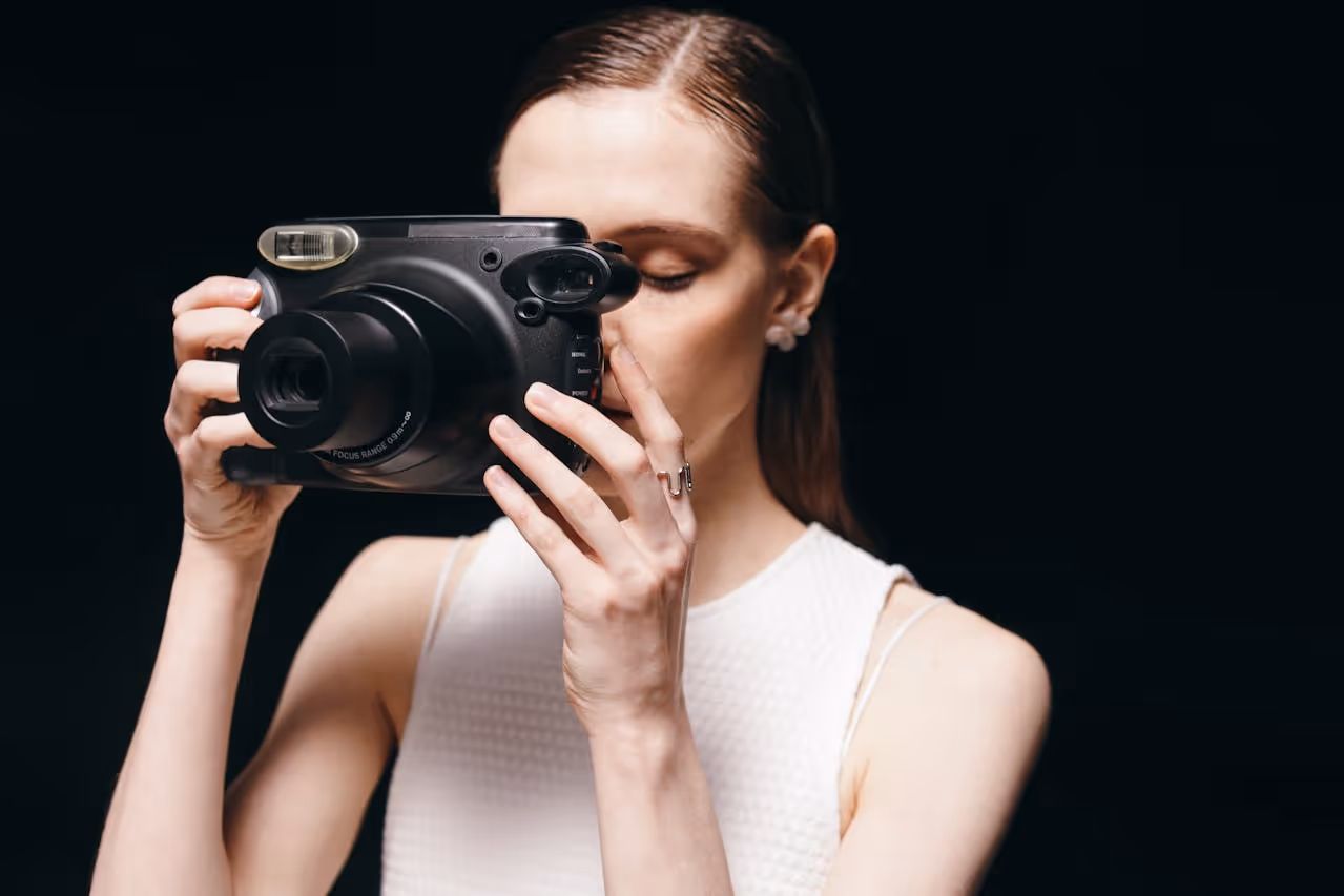 Woman in white sleeveless top holding a black instant camera close to her face against a dark background.