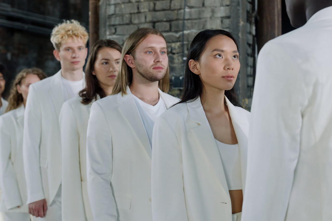 A diverse group of young adults dressed in white suits standing in a line indoors against a brick wall.