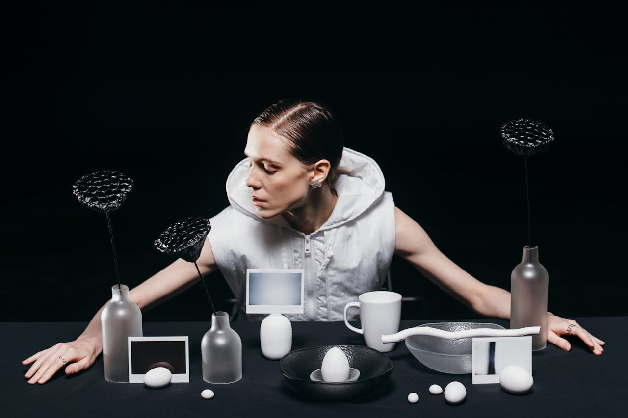 Woman in white sleeveless hoodie sitting behind a black table with frosted glass vases, black textured flowers, white eggs, a white mug, and artistic objects on a dark background.